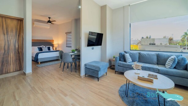 Una sala de estar moderna con sofá, mesa de centro y televisor. Se observa una cama al fondo y una vista a la ciudad a través de la ventana.