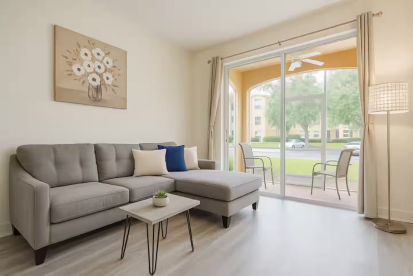 A modern living room with a grey sectional sofa, a small white coffee table, and a floor lamp. There is a large window with curtains and a view of a building and trees outside.
