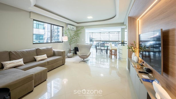 A modern living room with a sofa, a chair, a dining area, and a TV mounted on the wall. - Apt w/ 3 BR & Grill 400m From the Beach Prs0101 (Itapema)