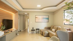 A modern living room with a flat-screen TV, a painting of a beach, and a window with a plant. - Apt w/ 3 BR & Grill 400m From the Beach Prs0101 (Itapema)