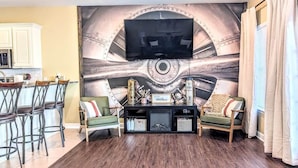 Een woonkamer met een grote muurschildering, een tv boven een zwarte entertainmentkast, twee groene fauteuils, een salontafel met decoratieve objecten, en een keukenhoek met witte kasten en een barkruk.