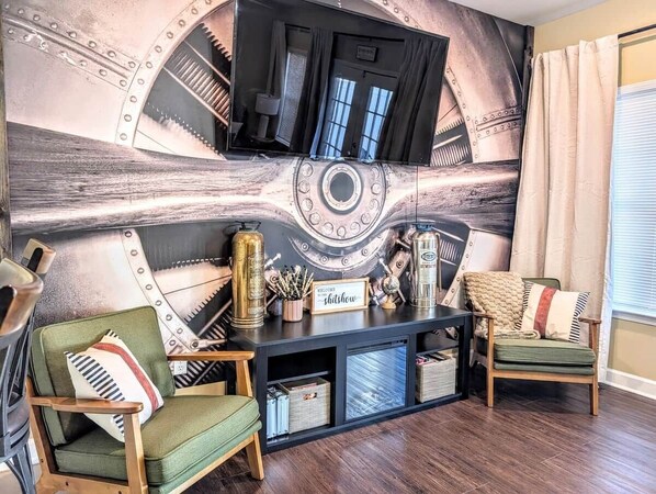 A living room with a large wall mural, two green armchairs, a wooden coffee table, and a television mounted on the wall.