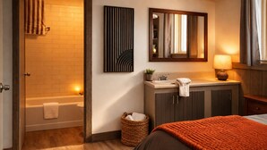 A hotel room with a wooden floor, a bed with an orange blanket, a bathroom with a bathtub, a sink, and a mirror.