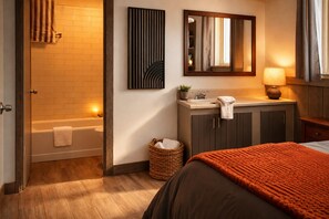 Una habitación de hotel con piso de madera, una cama con una manta naranja, un baño con bañera, lavamanos y espejo.