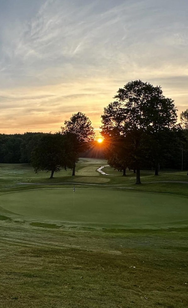 Campo de golf con un sendero despejado, árboles y un atardecer de fondo.