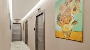 Un couloir avec trois portes, un tableau représentant des tournesols et un petit panneau en forme de maison accroché au mur.
