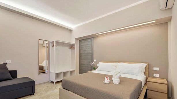 A modern hotel room with a bed, bedside tables, a wardrobe, a mirror, and a seating area.