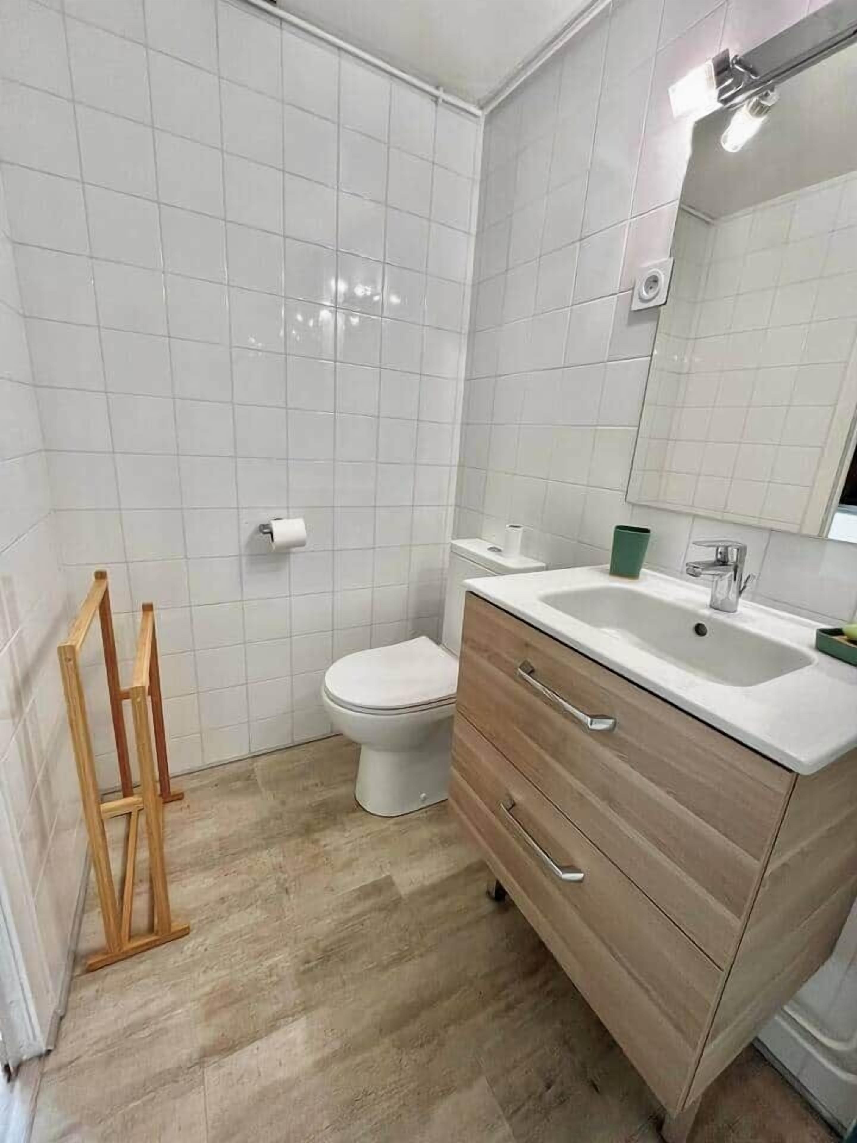 Een badkamer met witte tegels, een toilet, een houten wastafelmeubel met lades en een spiegel.