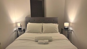 Un lit bien fait, avec du linge de lit blanc et deux oreillers, flanqué de deux tables de chevet surmontées de lampes, dans une pièce au design simple et épuré.