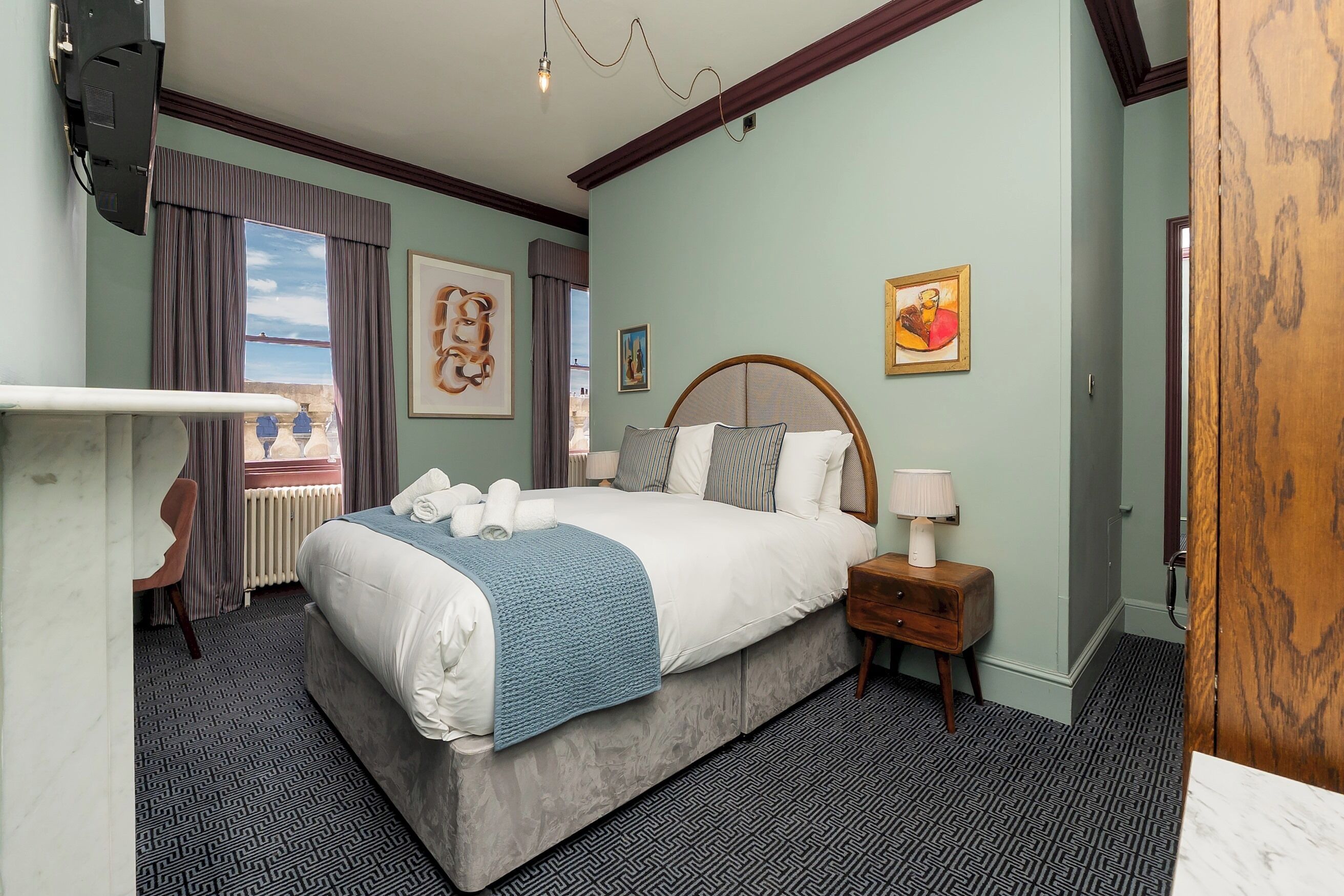 A hotel room with a bed, a desk, a chair, a nightstand, and a window with curtains.