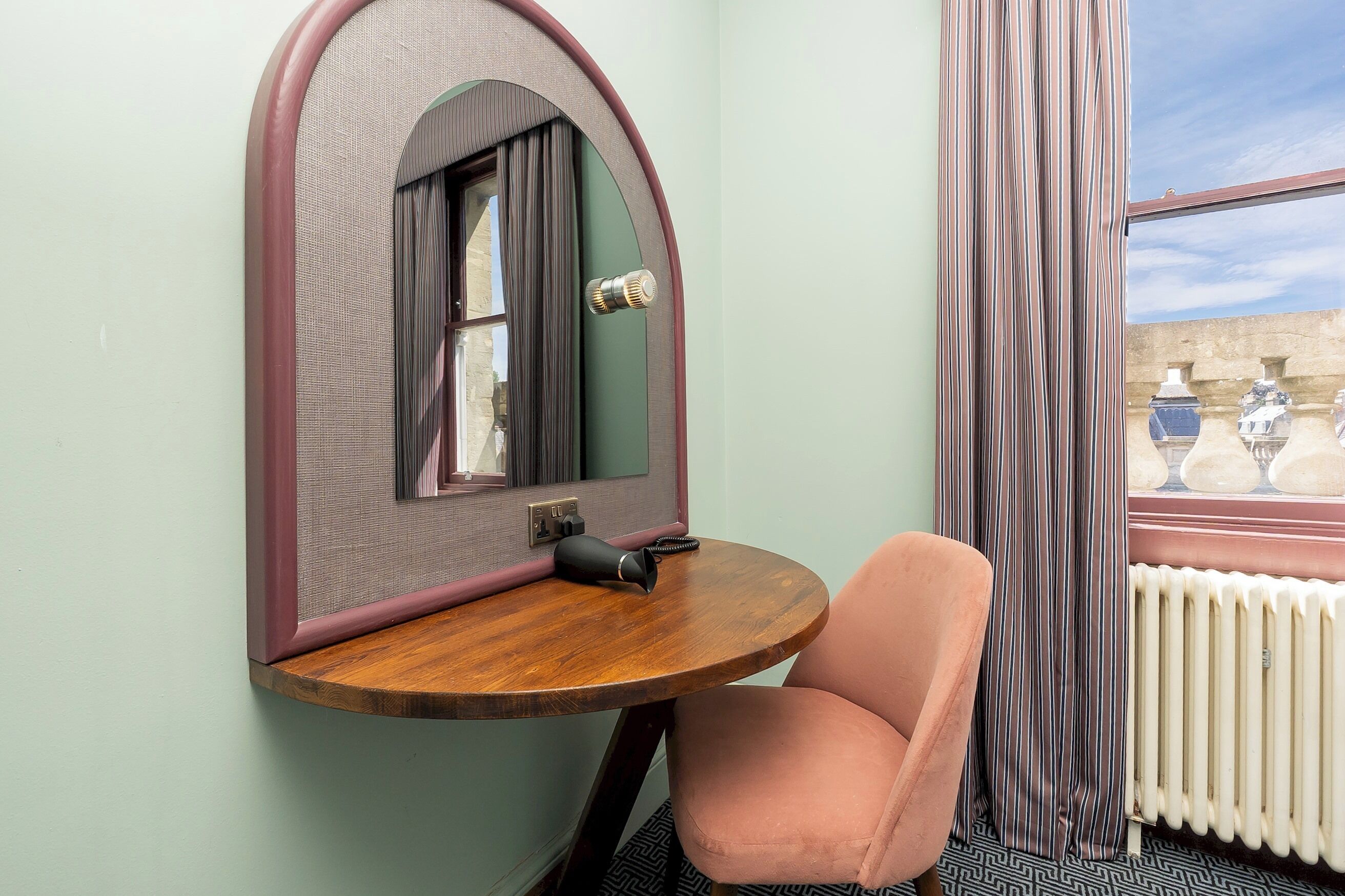 A compact room with a round wooden table, a single pink chair, a mirror with a unique arch design, and a window with curtains.