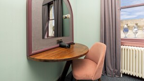 Een kleine kamer met een ronde houten tafel, een enkele roze stoel, een spiegel met een bijzonder boogvormig ontwerp, en een raam met gordijnen.
