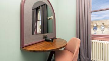 A compact room with a round wooden table, a single pink chair, a mirror with a unique arch design, and a window with curtains.
