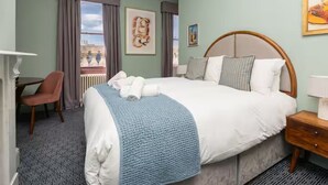 Une chambre d’hôtel avec un grand lit, un bureau et une chaise. Les fenêtres sont dotées de rideaux et les murs sont ornés d’œuvres d’art.