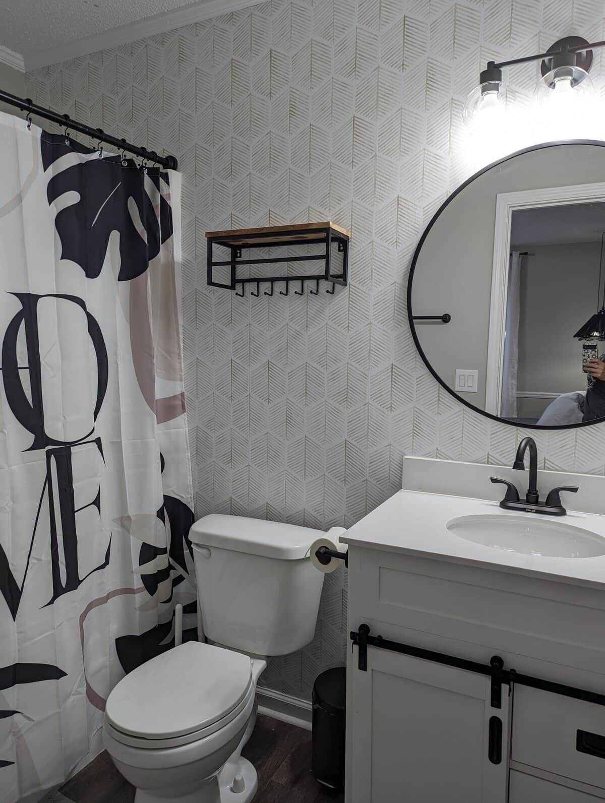 Une salle de bain avec des toilettes blanches, un lavabo avec un miroir et un rideau de douche orné d’un motif de feuilles.
