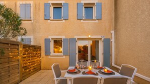An outdoor dining area with a table set for four, surrounded by white chairs, against a building with blue shutters and a wooden fence. - L’Échappée Belle, 4 guests, Pool in Calvisson (Calvisson)