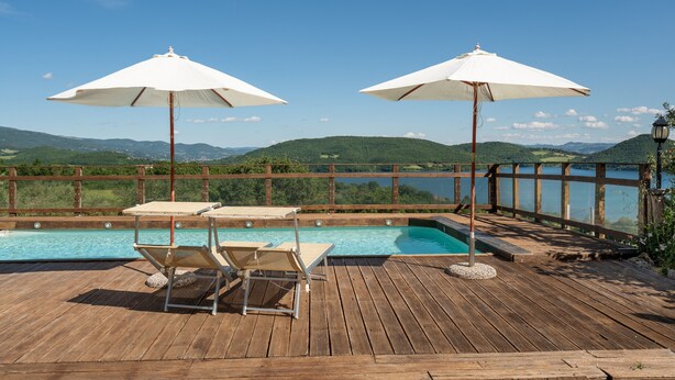 A wooden deck with a pool, two umbrellas, and lounge chairs.