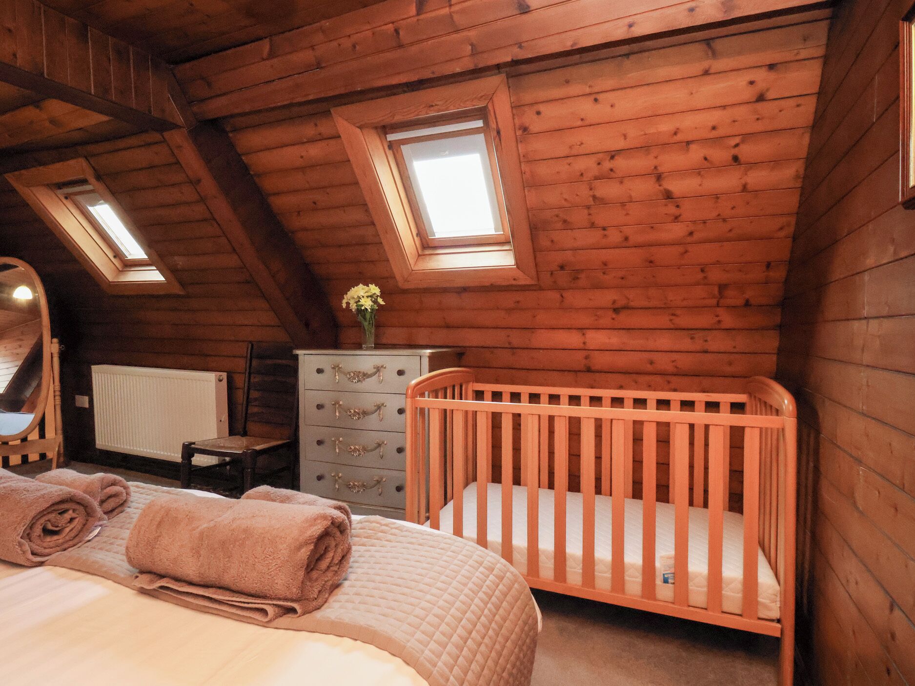 Ein gemütliches Schlafzimmer im Dachgeschoss mit schrägverdeckelter Holzbalkendecke, einem Wiege, einem Schminktisch und einem Dachfenster.