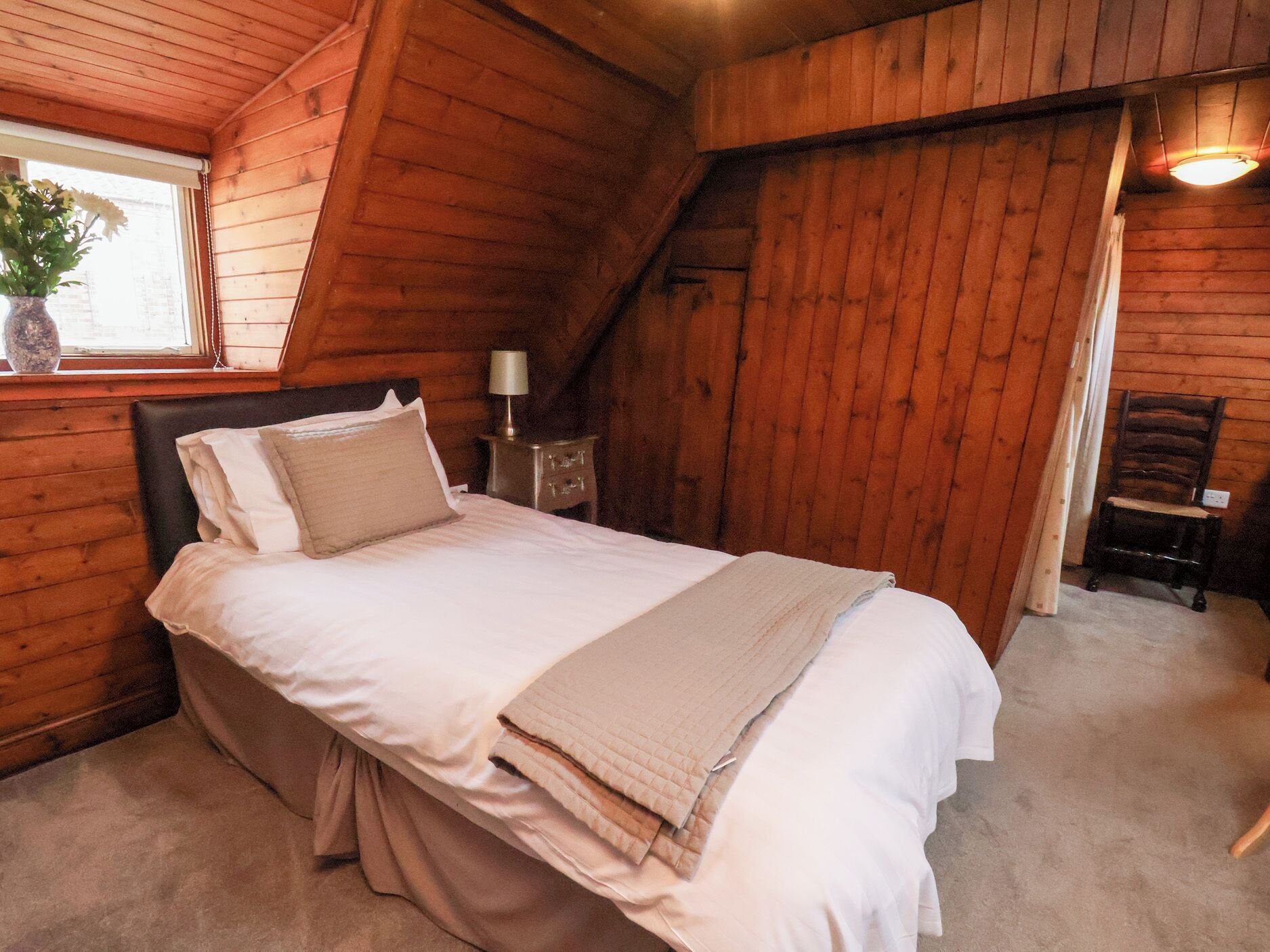 Una habitación acogedora de madera con una cama, una mesita de noche, una lámpara y una silla.