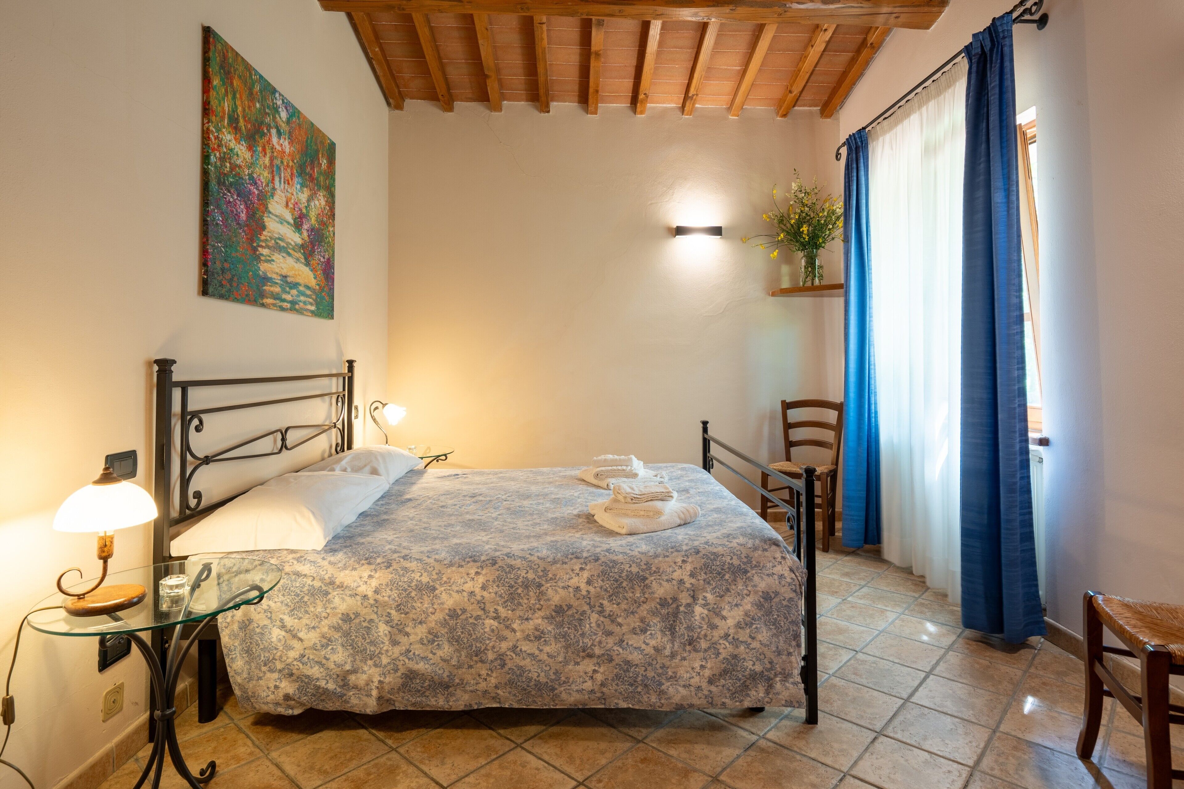 A bedroom with a bed, a nightstand, a lamp, a painting, a window with curtains, and a chair.