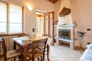 Un espace repas comprenant une table et des chaises, une cheminée et une vue sur l’extérieur, accessible par une porte ouverte.