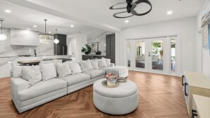 Un salon moderne avec un canapé d’angle gris, un pouf rond surmonté d’un vase, et un coin repas avec une table en bois.