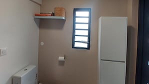 Casa de banho pequena e compacta com sanita, um prateleira em cima, uma janela e frigorífico.