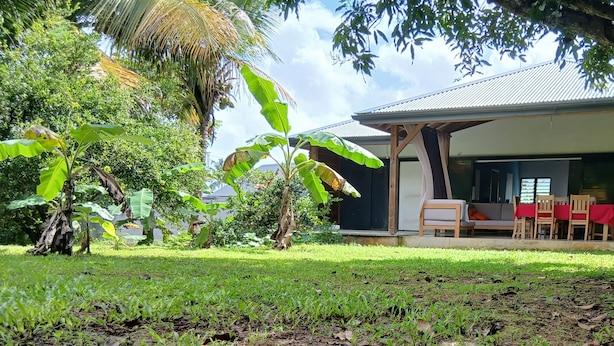 Uma área de estar coberta ao ar livre, com mesa e cadeiras, cercada por vegetação exuberante e uma bananeira.