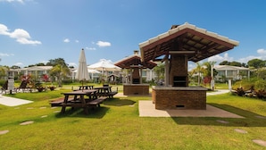 A patio area with picnic tables and a barbecue grill, surrounded by well-maintained grass and a clear sky. - House 2 Suites in Condo w/ Pool Mnt0058 (Porto Seguro)