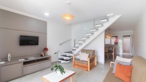 A modern living room with a flat-screen TV, a wooden console table, a staircase, and a dining area. - House 2 Suites in Condo w/ Pool Mnt0058 (Porto Seguro)