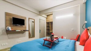 Quarto de hotel com uma cama, televisão presa à parede, uma mesinha com bandeja e uma cadeira vermelha.