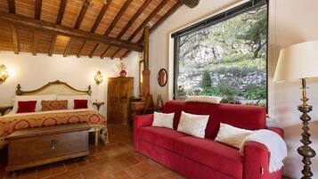Una stanza con un letto, un divano rosso, un comò in legno, una finestra con vista sul verde e un lampadario.