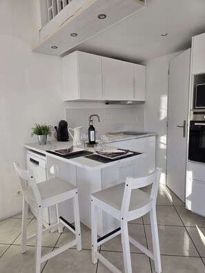 A compact kitchen with white cabinetry, a central island with seating, and a built-in oven. - Le Nid de Paulilles in Port-Vendres (Port-Vendres)