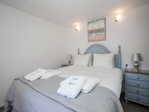 Un lit bien fait, avec du linge de lit blanc et deux oreillers, une couverture grise et deux serviettes pliées posées sur le lit. On y voit une tête de lit bleue, une table de chevet avec une lampe et un luminaire fixé au mur.