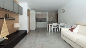 A modern living room with a sofa, a coffee table, and a TV. There is a dining area with a table and chairs. The kitchen is visible in the background with cabinets and a countertop. - Zardin 7 - One Bedroom Apartment, Sleeps 4 (Valdidentro)