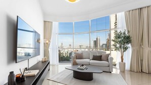 A modern living room with a large window, a flat-screen TV, a sofa, a coffee table, and a potted plant. - Stayshort - Stylish 1-bedroom Home Fully Furnished in JVC (Dubai)