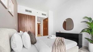 A modern bedroom with a bed, a dresser, a mirror, and a potted plant. - Stayshort - Stylish 1-bedroom Home Fully Furnished in JVC (Dubai)