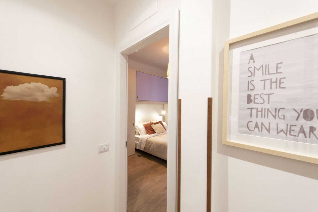 A hallway with a framed quote on the wall, a painting of a cloud, and a glimpse of a bedroom with a bed and a purple wall.