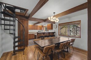 A modern kitchen with wooden cabinets, a dining table with chairs, a spiral staircase, and a large window with a view. - Modern Sunriver Retreat – Sleeps 16 (Sunriver)