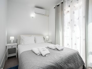 A hotel room with a bed, two bedside tables with lamps, a wall-mounted air conditioner, and a window with sheer curtains. - Dappan White Apartment, Burgau, Algarve (Burgau)