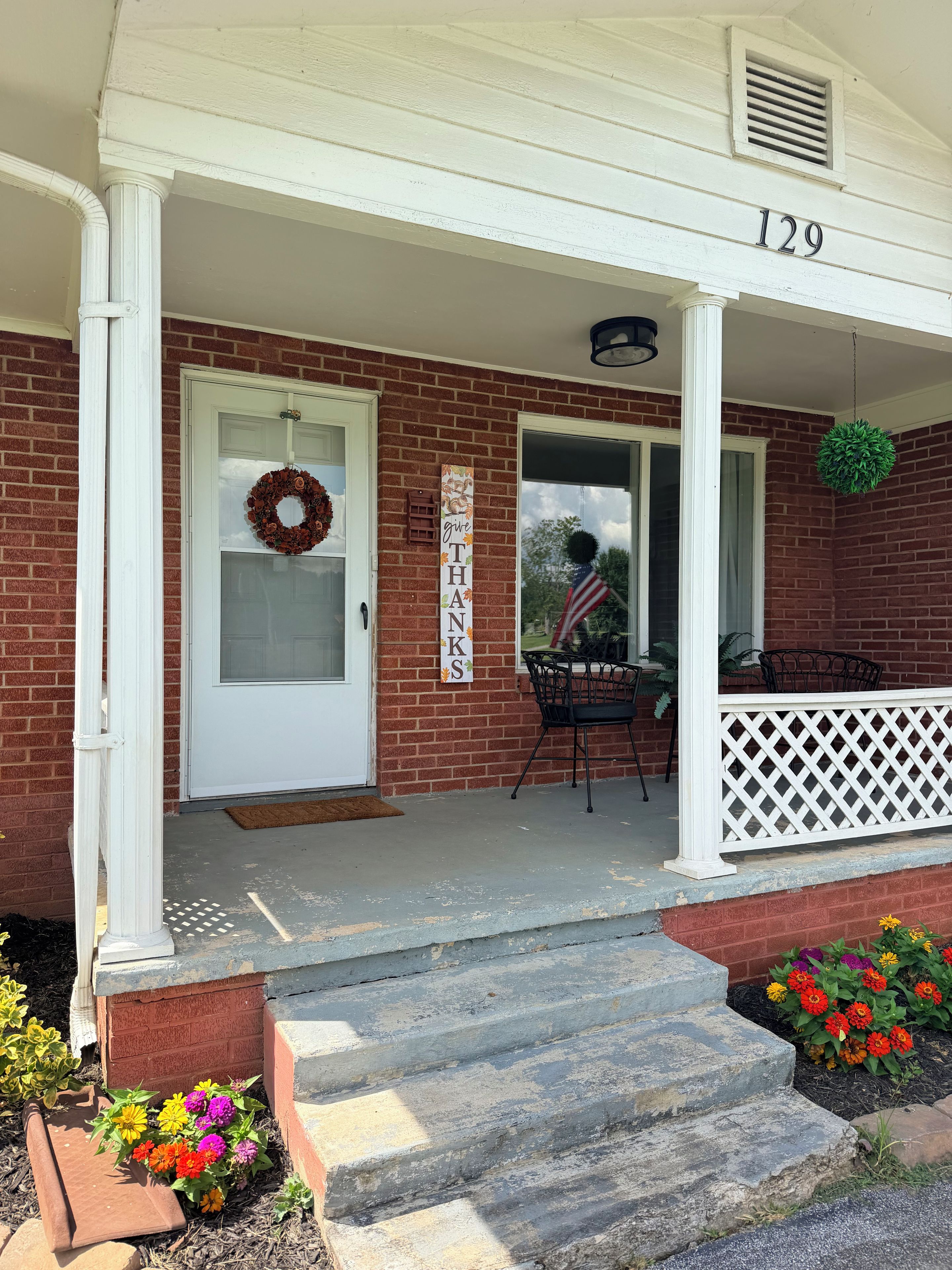 A brick house with a white porch, a door with a wreath, and a sign that says 'Give Thanks.'