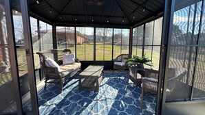 Eine überdachte Terrasse mit Korbmöbeln, einem gemusterten Teppich und Blick auf einen Rasen und Gebäude.