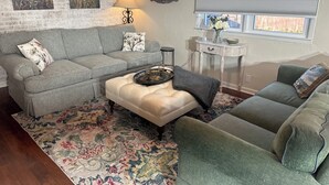 Ein Wohnzimmer mit einem grauen Sofa, einem cremefarbenen Ottomanen und einem grünen Sessel. Auf dem Boden liegt ein gemustertes Teppich.