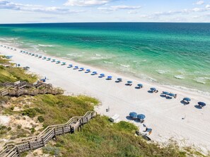 Ein Strand mit Reihen blauer Sonnenschirme und Liegestühle, Holzstege und freiem Blick auf das Meer.