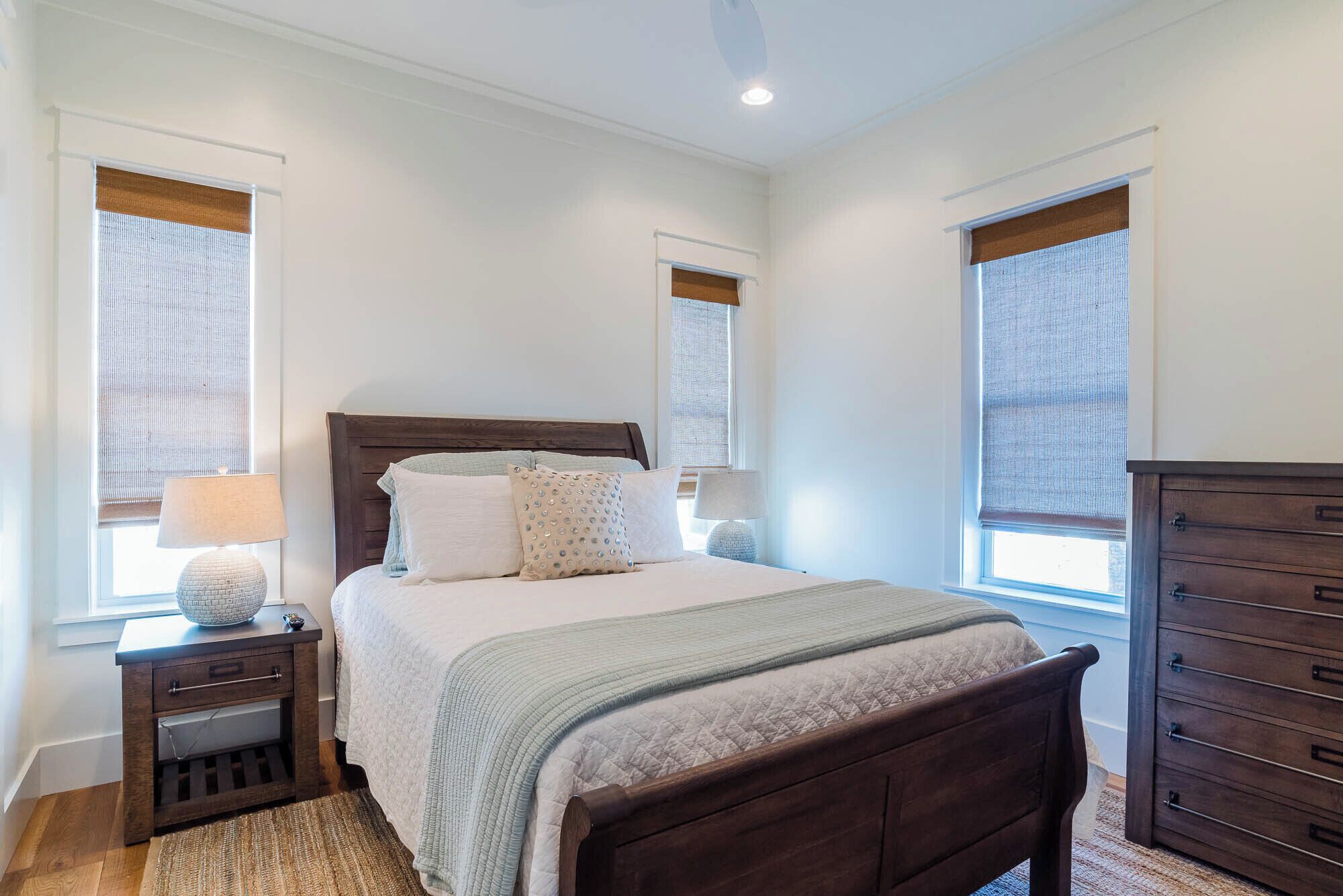 A bedroom with a wooden bed, two bedside tables with lamps, a dresser, and large windows with blinds.