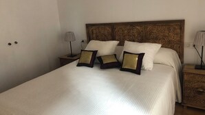 Ein ordentlich bezogenes Bett mit weißer Decke und drei Zierkissen, ein Holz-Kopfschild mit aufwendigen Schnitzereien sowie zwei Nachttische mit Lampen.