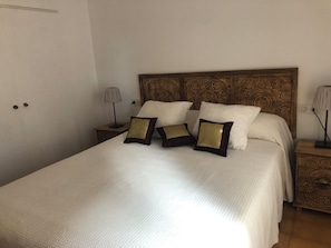 Ein ordentlich bezogenes Bett mit weißer Decke und drei Zierkissen, ein Holz-Kopfschild mit aufwendigen Schnitzereien sowie zwei Nachttische mit Lampen.