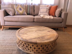 Ein beiges Sofa mit gemusterten Kissen und einem runden Couchtisch aus Holz mit Rautenmuster.