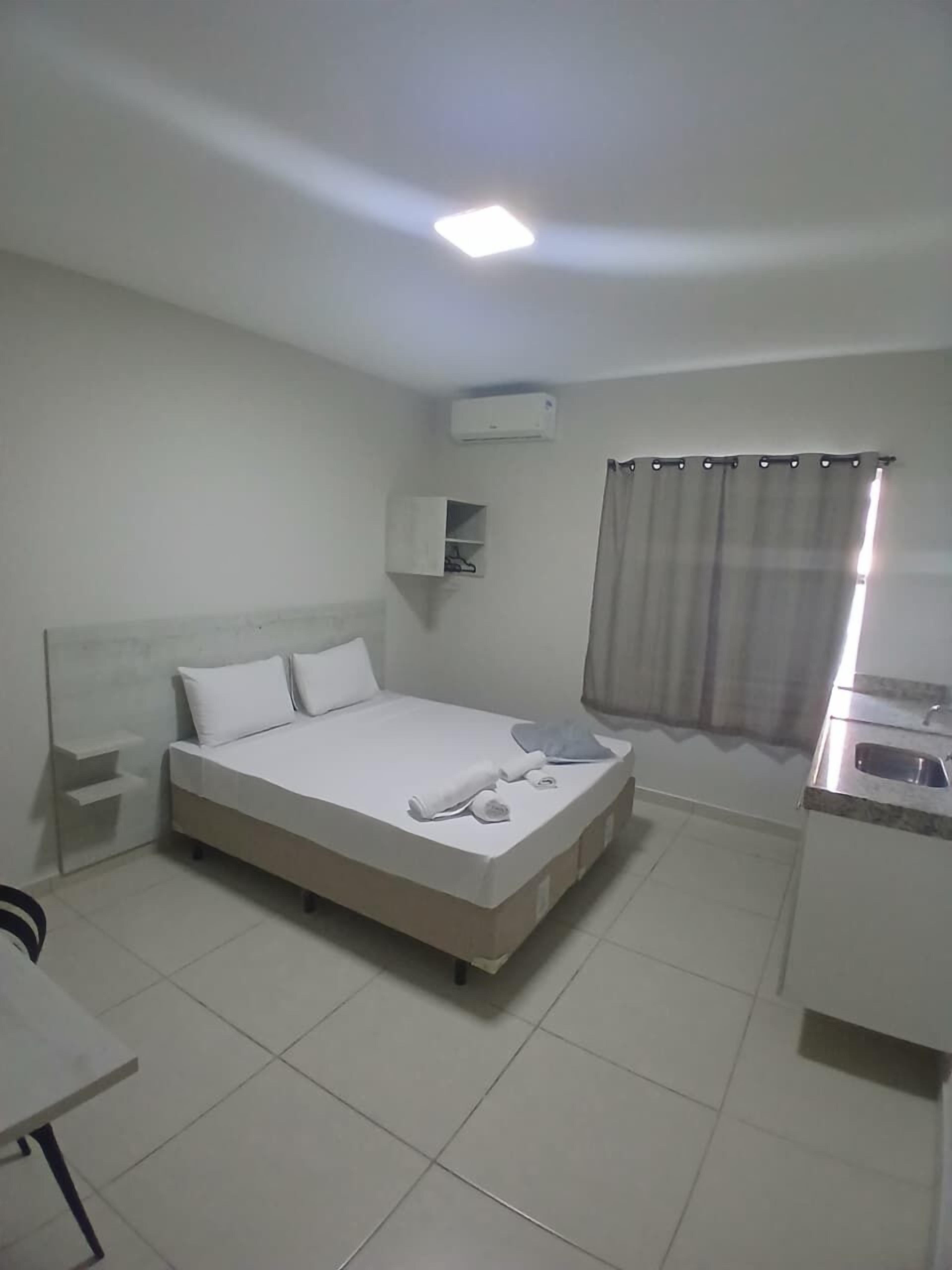 Habitación de hotel con cama, mesita de noche, aire acondicionado integrado, ventana con cortina y kitchenette con lavabo.