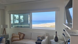 Ein Zimmer mit großem Fenster mit Blick auf einen Strand, ein weißes Sofa, eine Lampe und ein kleiner Tisch mit einer Vase.